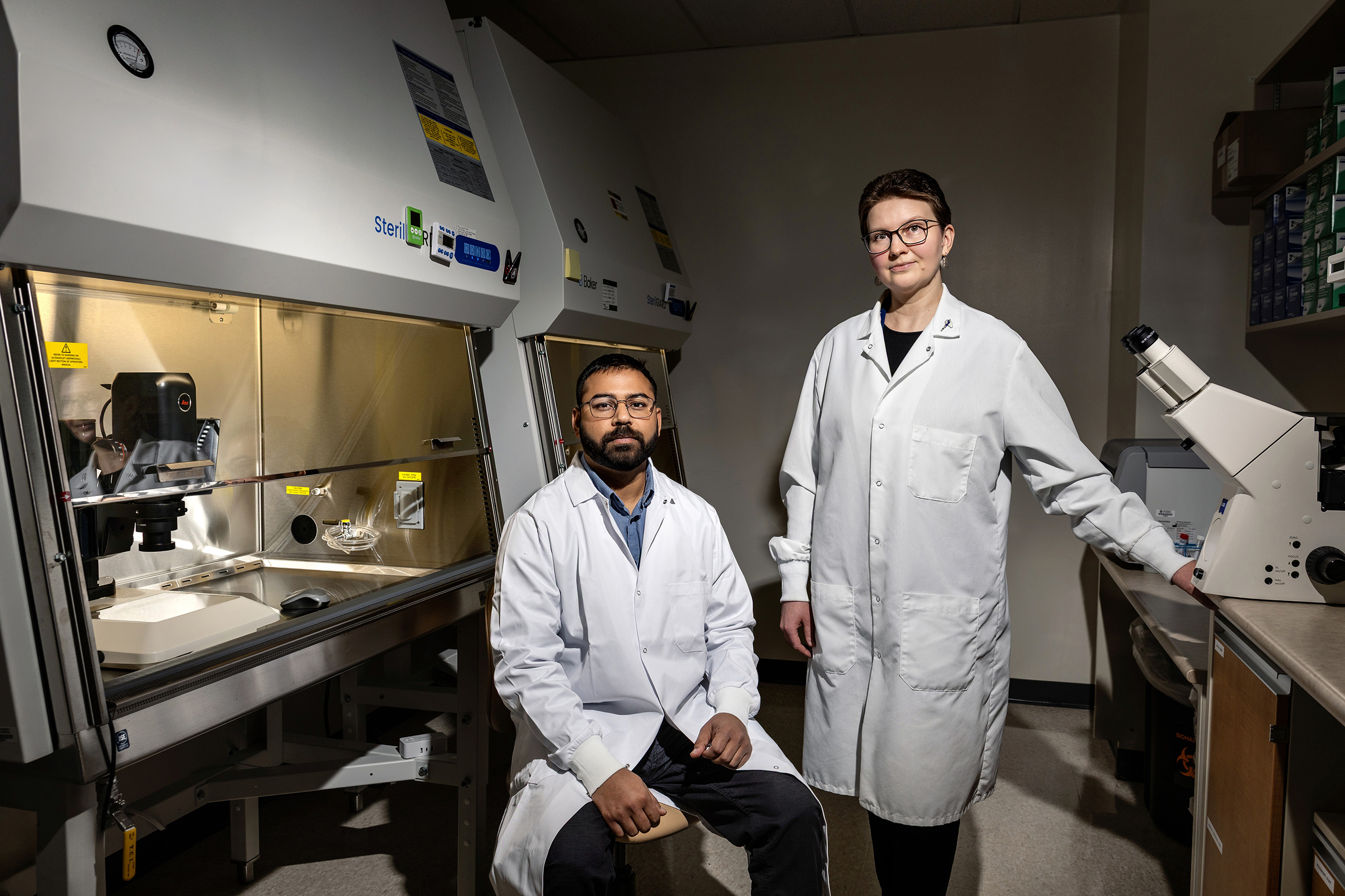 Two people wearing lab coats and serious expressions in a small lab filled with scientific equipment